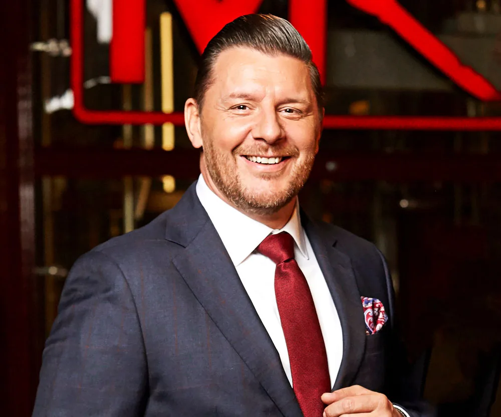 Man smiling, wearing a suit and red tie, with "MKR" logo blurred in background.