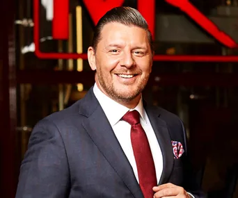 Man smiling, wearing a suit and red tie, with "MKR" logo blurred in background.