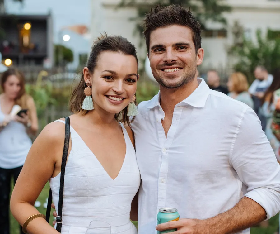 A smiling couple in white outfits, enjoying a casual outdoor gathering.