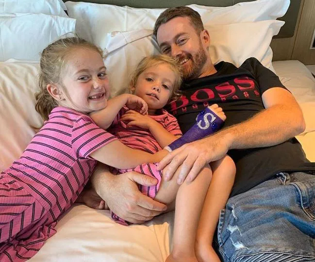 Father with an injured arm rests on a bed, smiling with his two young daughters in matching pink outfits.