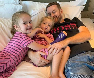 Father with an injured arm rests on a bed, smiling with his two young daughters in matching pink outfits.