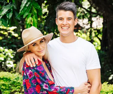 A smiling couple poses outdoors, the woman in a floral top and hat, the man in a white shirt, surrounded by greenery.