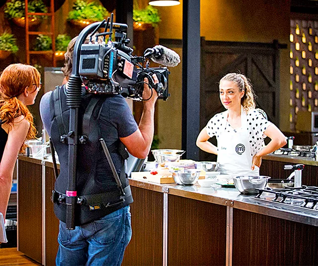 Cameraman films a MasterChef contestant in a kitchen set, with cookware on table, bright lighting and greenery decor.