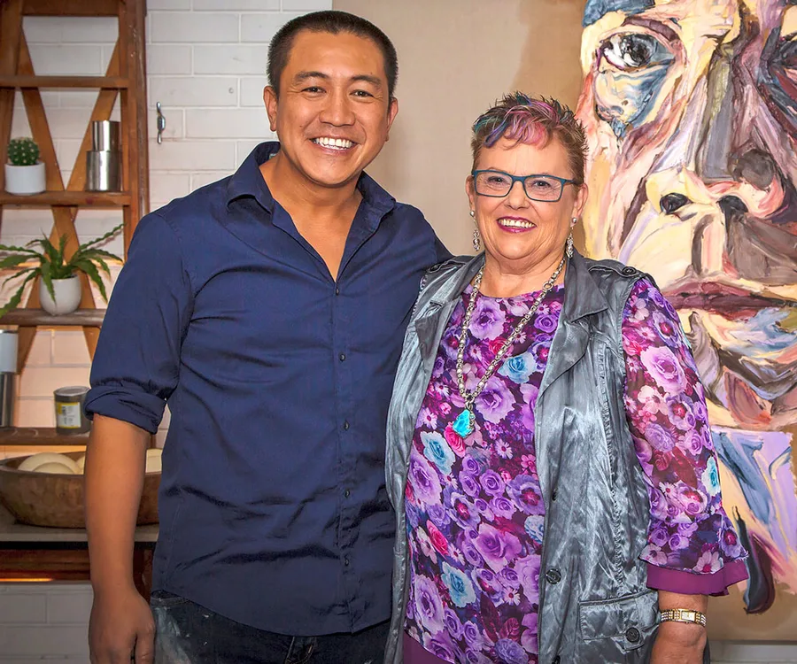 Two people smiling, standing together indoors near a colorful painting in a studio setting.