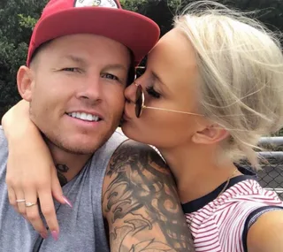 A woman kisses a man on the cheek outdoors; he wears a red cap, and she has sunglasses and a tattooed arm.