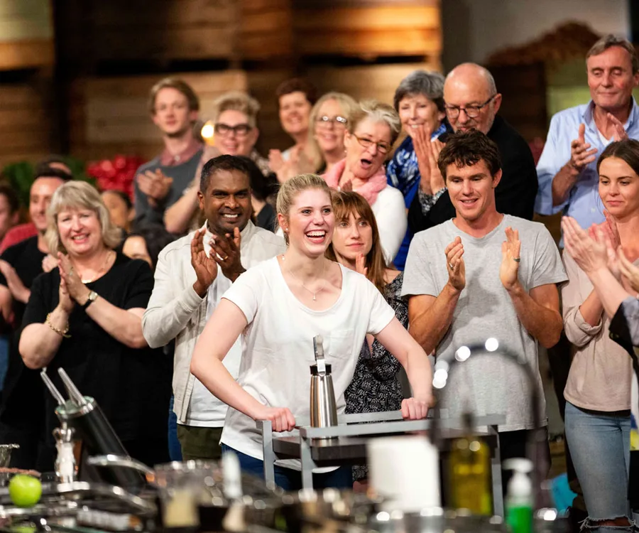 "MasterChef Australia contestant in a white shirt smiles while a crowd applauds in the background."
