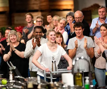 "MasterChef Australia contestant in a white shirt smiles while a crowd applauds in the background."