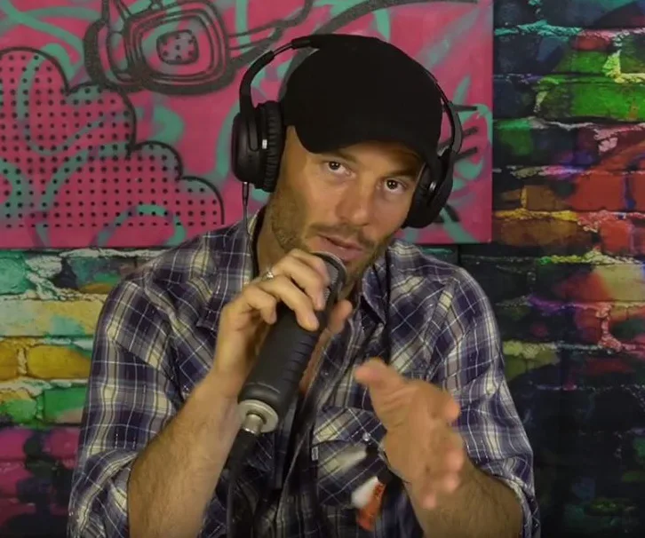Man in a black cap with headphones, holding a microphone, in front of a colorful graffiti-style backdrop.