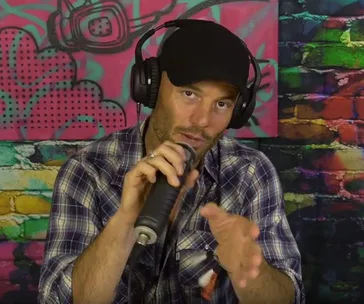 Man in a black cap with headphones, holding a microphone, in front of a colorful graffiti-style backdrop.