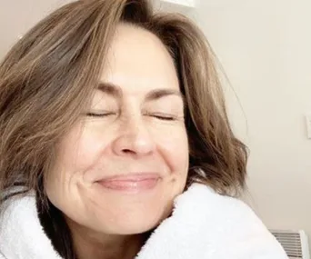 A woman with brown hair smiling, eyes closed, wearing a white robe, with a calm expression.