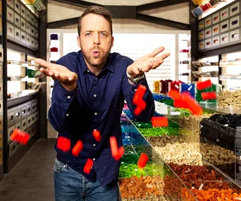 Host in a blue shirt stands in a LEGO store, playfully tossing red LEGO bricks with shelves of colorful bricks behind.
