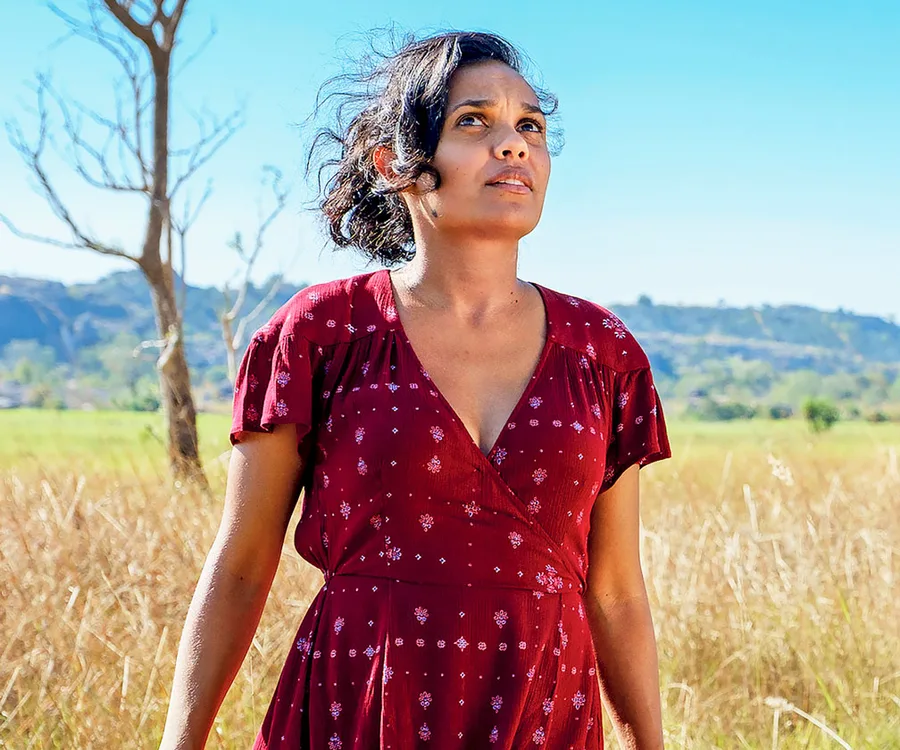 A woman in a red dress stands in a field under a clear blue sky, looking thoughtfully into the distance.