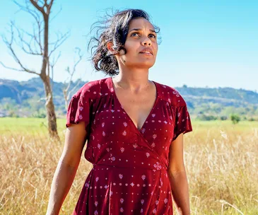 A woman in a red dress stands in a field under a clear blue sky, looking thoughtfully into the distance.