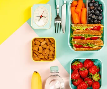 Healthy lunchbox with sandwiches, strawberries, almonds, carrots, blueberries, and a banana on a colorful background.