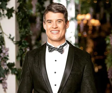 Man smiling in a tuxedo in front of a floral backdrop.