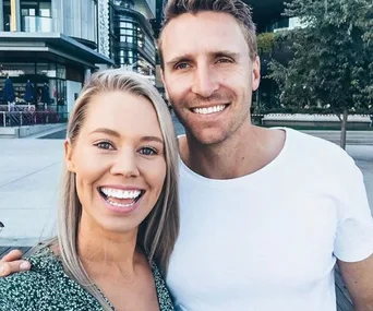 A smiling couple taking a selfie outdoors, with modern buildings and trees in the background.
