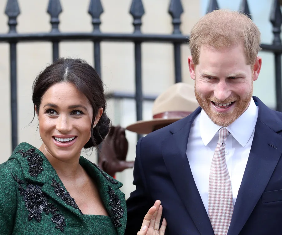 Two smiling individuals, one in a green outfit and the other in a suit, stand outdoors near a black fence.