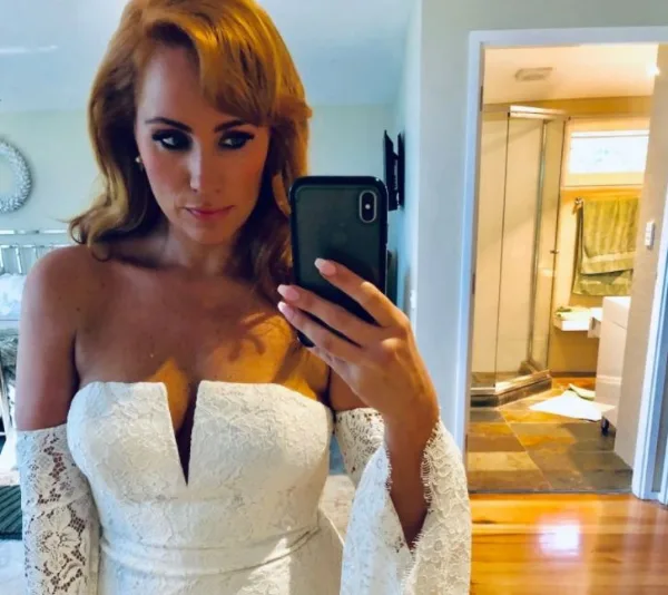 Woman taking a selfie in a mirror wearing an off-shoulder lace wedding dress.