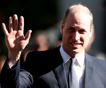 A person in a suit waves with a smile in bright sunlight.