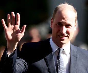 A person in a suit waves with a smile in bright sunlight.