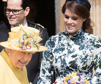 Two women smiling; one in a yellow hat with flowers, the other in a floral dress, holding a bouquet.