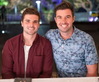 Two men sitting side by side, smiling at the camera, one in a maroon shirt and the other in a palm tree patterned shirt.
