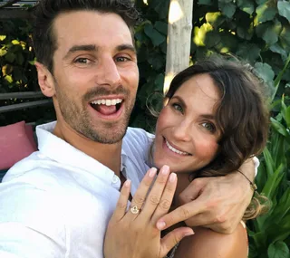 A couple smiling outdoors, with the woman showing an engagement ring.