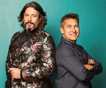 Two men pose confidently against a teal background; one in a patterned suit, the other in a dark jacket.