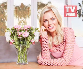 Blonde woman in a pink dress smiling beside a vase of pink and white flowers, with TV WEEK logo in the background.