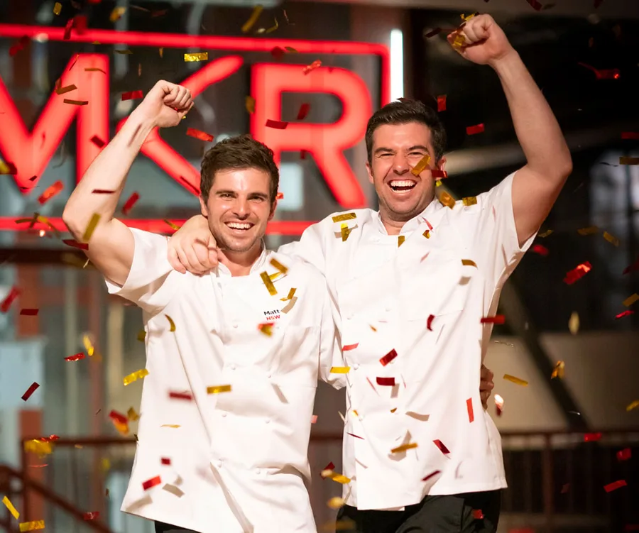Two chefs celebrating with confetti and a bright red "MKR" sign in the background, both in white uniforms.