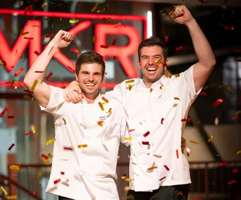 Two chefs celebrating with confetti and a bright red "MKR" sign in the background, both in white uniforms.