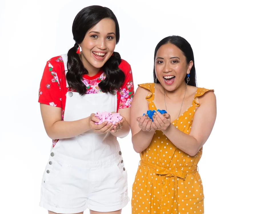 Two women smiling, holding pink and blue LEGO pieces, dressed in colorful outfits against a white background.