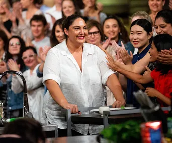 Person in white shirt receiving applause from crowd on MasterChef set.