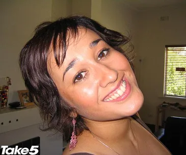 Smiling woman with short dark hair and pink earrings in a room with a window and furniture.