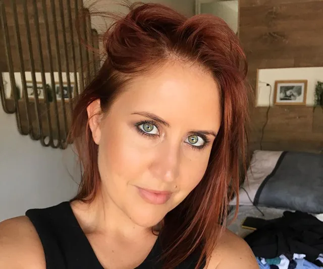 Red-haired woman with green eyes smiling in a bedroom, wearing a black top.