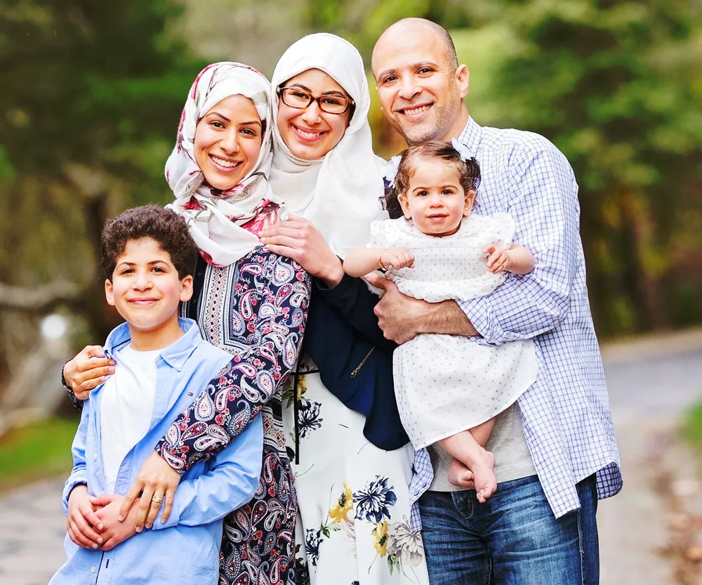 A family of five, including two women in hijabs, a young boy, a man, and a baby, smiling outdoors in a park setting.