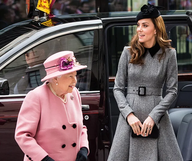 Two women smiling and walking near a car, one in a pink outfit and hat, the other in a gray coat and black hat.