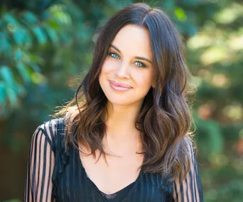 A woman with long brown hair and a sheer black top smiles at the camera, against a blurred green background.