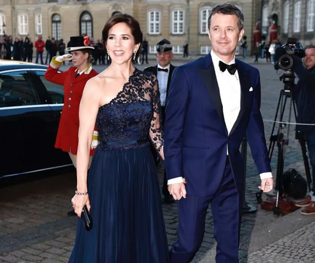 Crown Princess Mary and a man in formal attire at an outdoor event, with photographers and guards in the background.