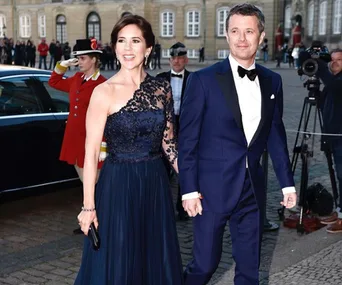 Crown Princess Mary and a man in formal attire at an outdoor event, with photographers and guards in the background.
