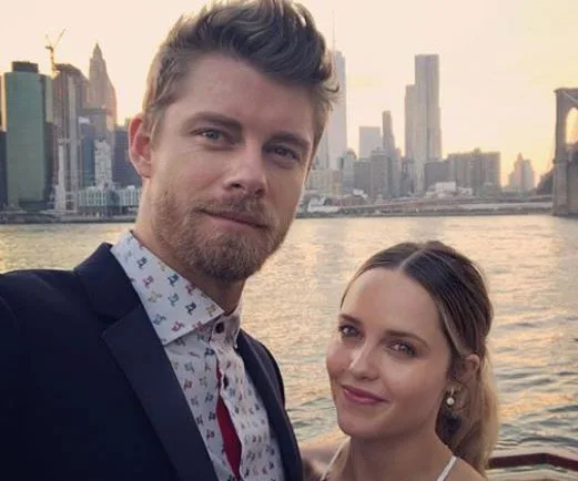 Man and woman smiling with New York City skyline and water in the background.