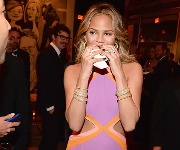 A woman in a colorful dress holding a napkin to her mouth at a social event with people in the background.