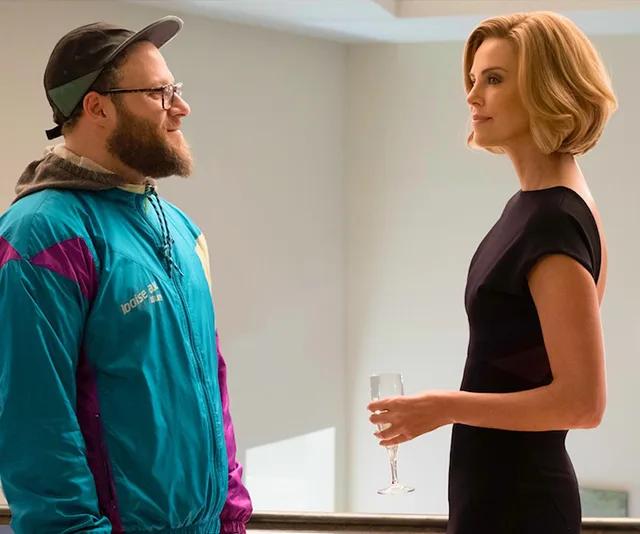 Two people standing and conversing; one in a colorful jacket, the other in a black dress holding a glass.