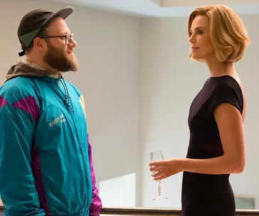 Two people standing and conversing; one in a colorful jacket, the other in a black dress holding a glass.