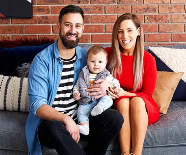 Family sitting on a couch, man wearing denim, woman in red dress, holding a baby wearing a vest and striped shirt.