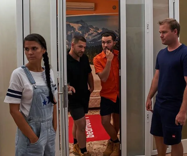 Four people inside a room: a woman in overalls with a braid and three men, two standing in a doorway, one in blue nearby.