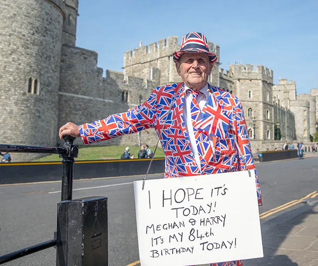 Man in Union Jack suit holds a sign about Meghan and Harry's baby outside a castle, coinciding with his 84th birthday.