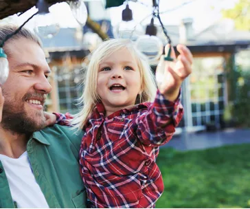 Father holding a happy toddler reaching for string lights outdoors.