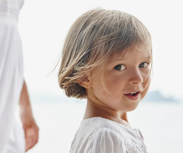 Toddler with short hair in a white dress smiles at the camera on a bright day near water.