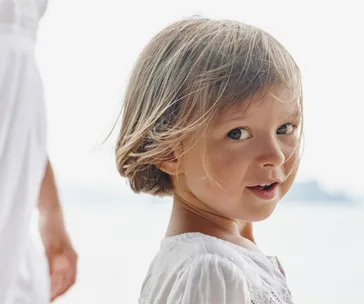 Toddler with short hair in a white dress smiles at the camera on a bright day near water.